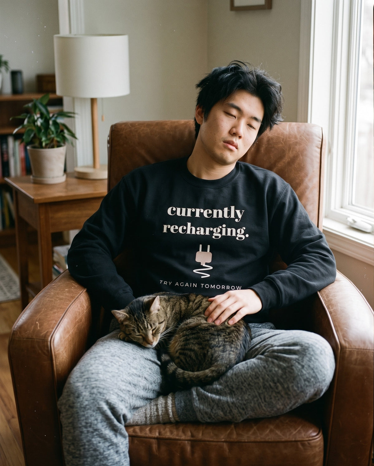 Currently Recharging Crewneck in Black, worn by a man dozing in a leather armchair with a tabby cat asleep on his lap. Printed on the chest: currently recharging, a plug and TRY AGAIN TOMORROW.