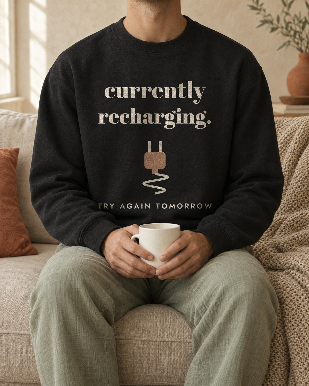 Currently Recharging Crewneck in Black, worn on a sofa with a mug in both hands. Printed on the chest: currently recharging, a plug icon and the line try again tomorrow.
