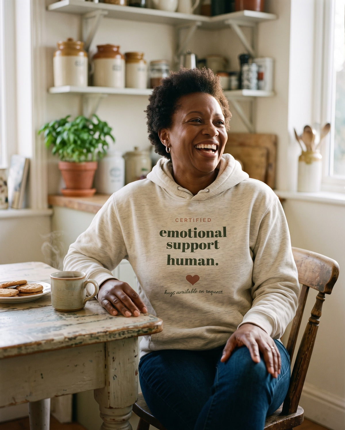 Emotional Support Human hoodie in Oatmeal Heather, worn by a woman laughing at a kitchen table with tea and biscuits. Printed on the chest: CERTIFIED emotional support human., a heart and the line hugs available on request.