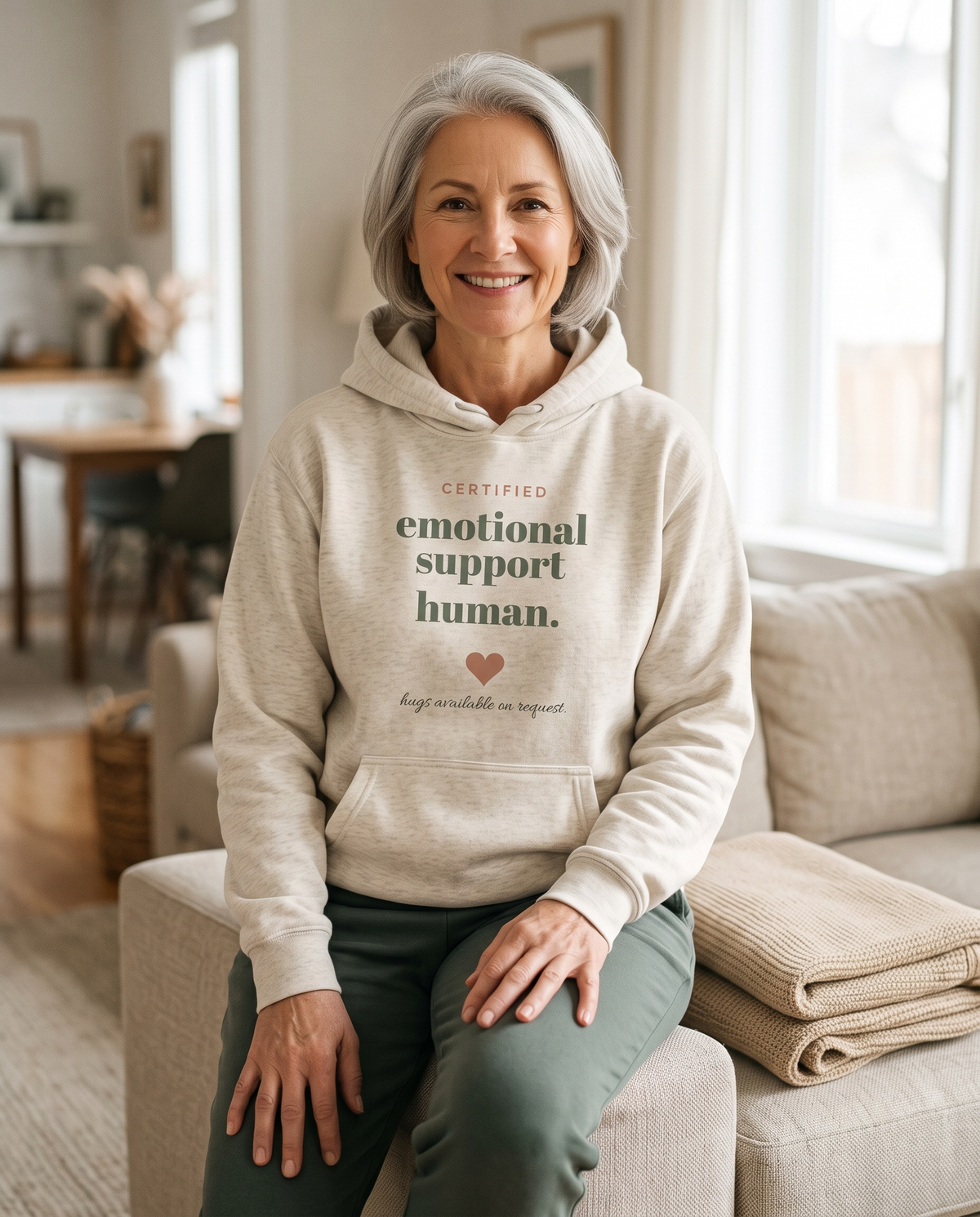 Emotional Support Human hoodie in Oatmeal Heather, worn by a smiling model sitting on the arm of a sofa. Printed on the chest: certified emotional support human, with a heart and hugs available on request.
