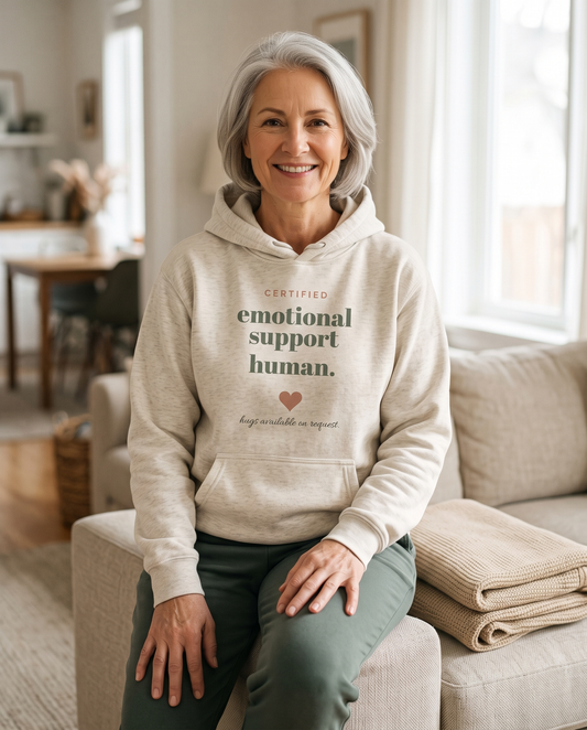 Emotional Support Human hoodie in Oatmeal Heather, worn by a smiling model sitting on the arm of a sofa. Printed on the chest: certified emotional support human, with a heart and hugs available on request.