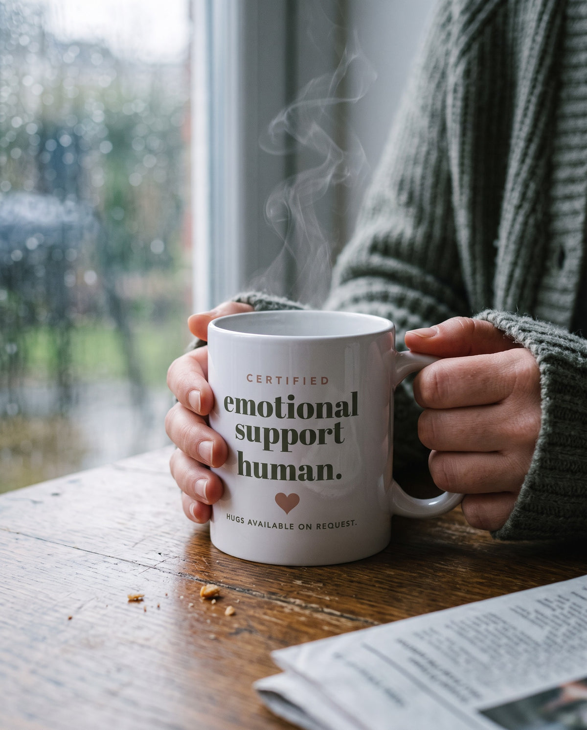 Emotional Support Human Mug, held at a kitchen table by a rainy window