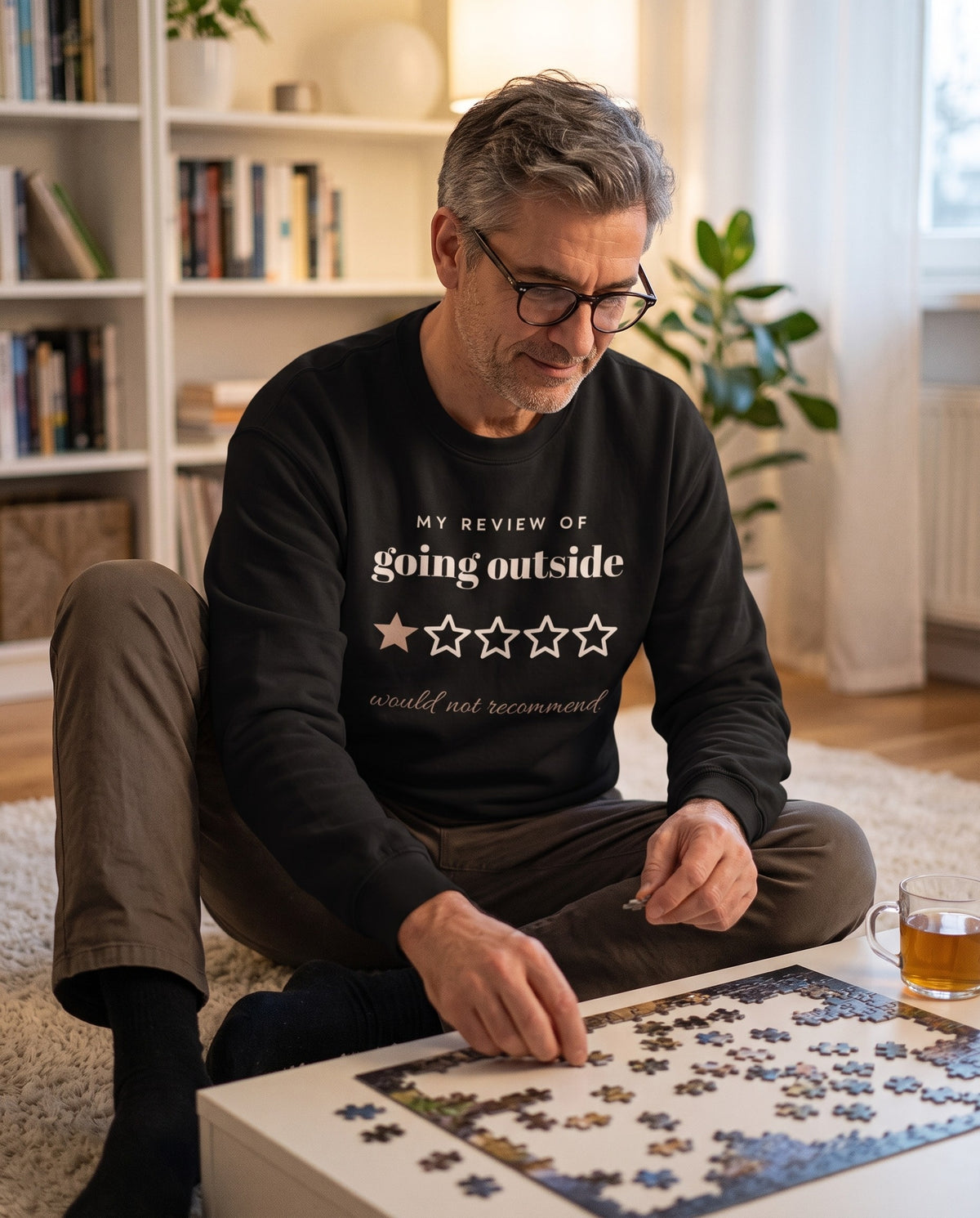 Going Outside, One Star crewneck in Black, worn by a man doing a jigsaw puzzle on the living room floor. Printed on the chest: MY REVIEW OF going outside, one star out of five and the line would not recommend.