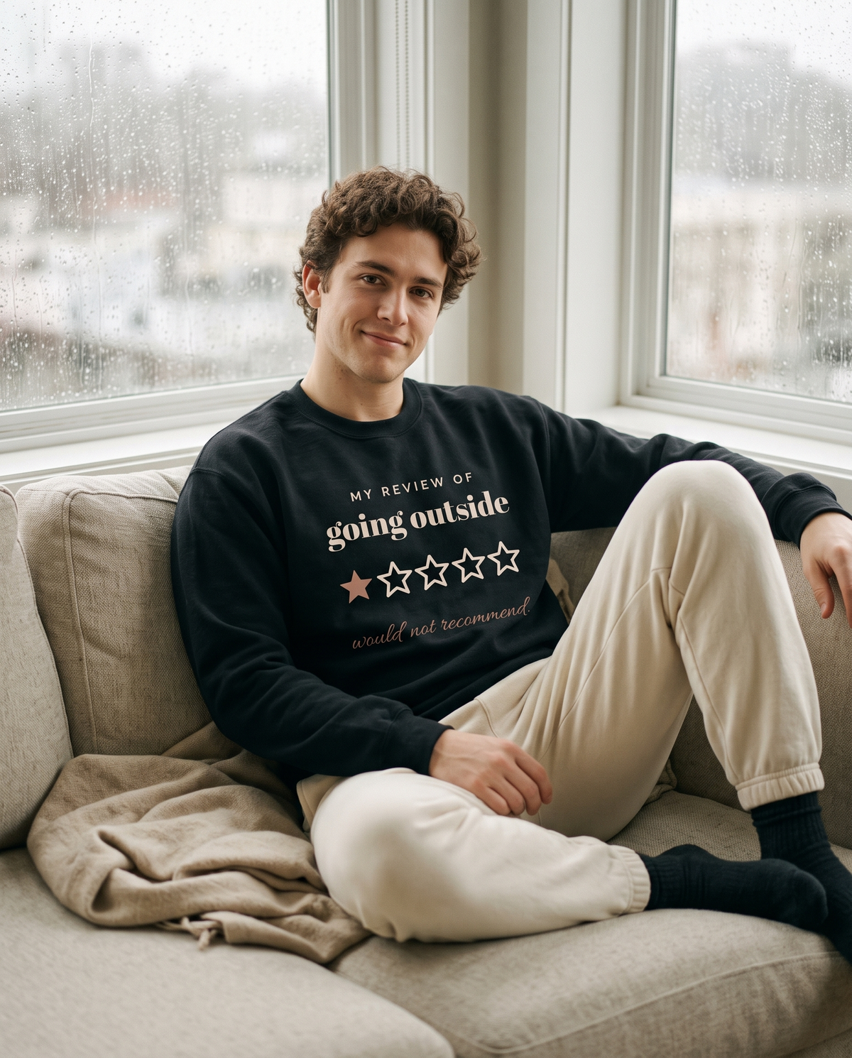 Going Outside, One Star crewneck in Black, worn by a model on a sofa by a rainy window. Printed on the chest: my review of going outside, one star out of five, would not recommend.