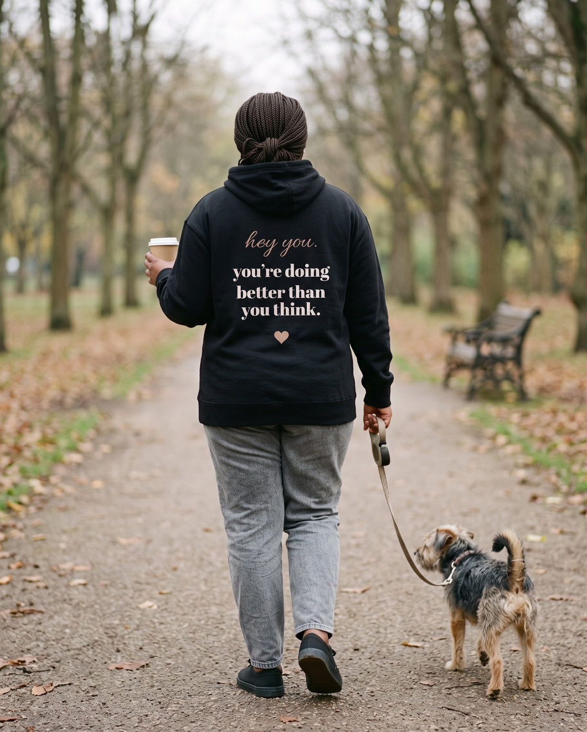Hey You Hoodie in Black, seen from behind on an autumn walk with a takeaway cup and a small dog. Printed on the back: "hey you. you're doing better than you think." with a small heart.