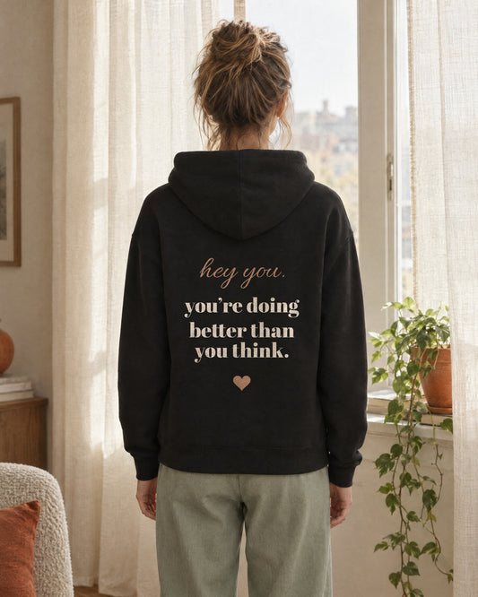 Hey You Hoodie in Black, worn by a model at a sunny window, seen from behind. The back print reads hey you, you are doing better than you think, with a small heart.