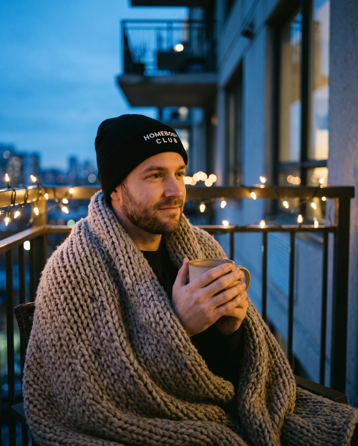 Homebody Club Beanie in Black, worn by a man wrapped in a knitted blanket on a balcony at dusk with a mug. Embroidered on the cuff: HOMEBODY CLUB.