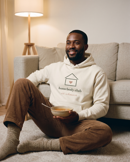 Homebody Club hoodie in Bone, worn by a model sitting on the living room floor against a sofa with a warm bowl. Printed on the chest: homebody club, est. whenever, with a small house and a heart.