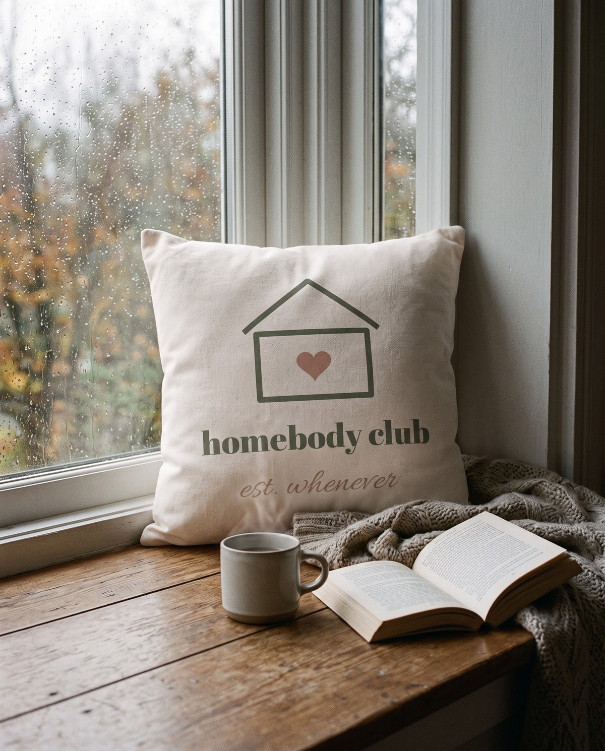 Homebody Club Pillow, on a window seat on a rainy day