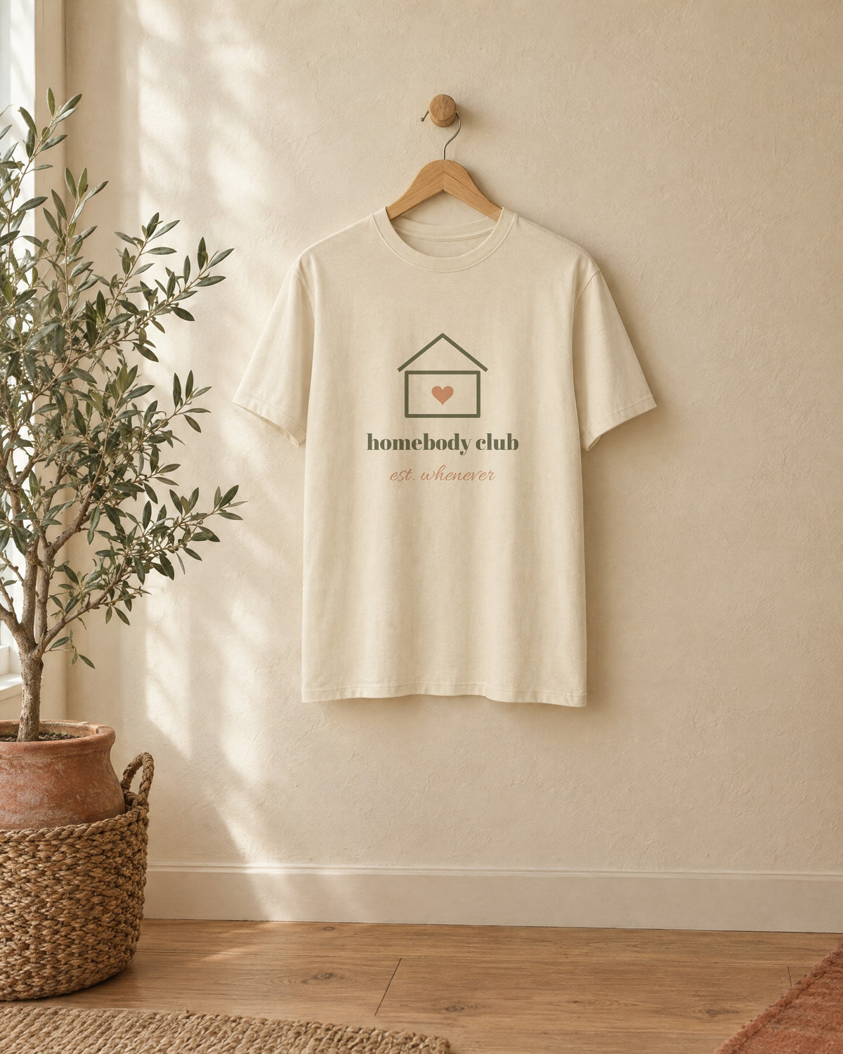 Homebody Club Tee, in soft cream, on a wooden hanger against a cream wall