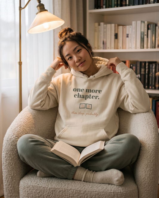 One More Chapter hoodie in Bone, worn by a model curled up in an armchair with a book. Printed on the chest: one more chapter, with an open book and then sleep, probably.