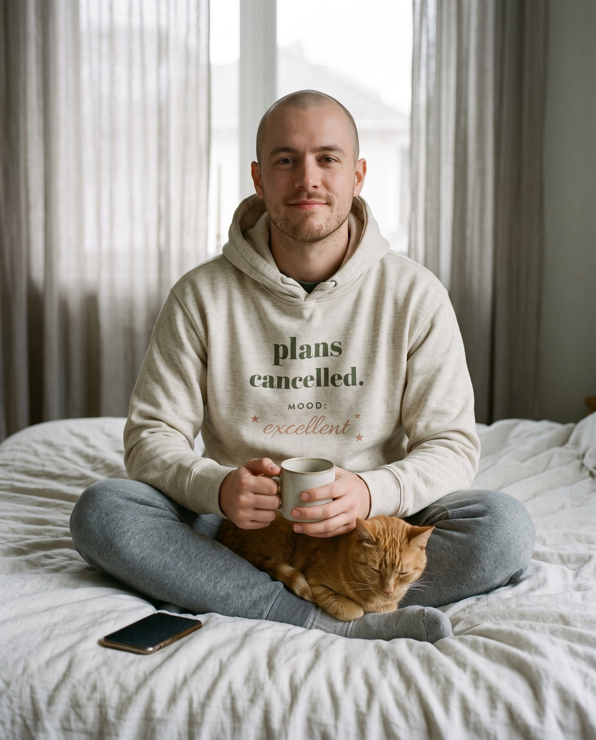 Plans Cancelled hoodie in Oatmeal Heather, worn by a man sitting on an unmade bed with a mug and a sleeping ginger cat. Printed on the chest: plans cancelled. MOOD: excellent, with small stars.