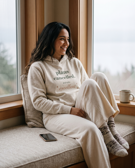 Plans Cancelled hoodie in Oatmeal Heather, worn by a model smiling on a window seat. Printed on the chest: plans cancelled, mood: excellent.