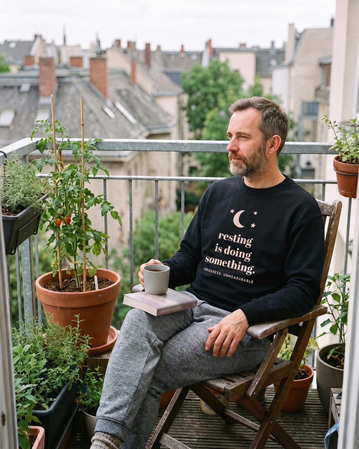Resting Is Doing Something crewneck in Black, worn by a man with a mug of tea on a city balcony full of plants. Printed on the chest: a moon and three stars, resting is doing something. and the small line PROGRESS: UNMEASURABLE.