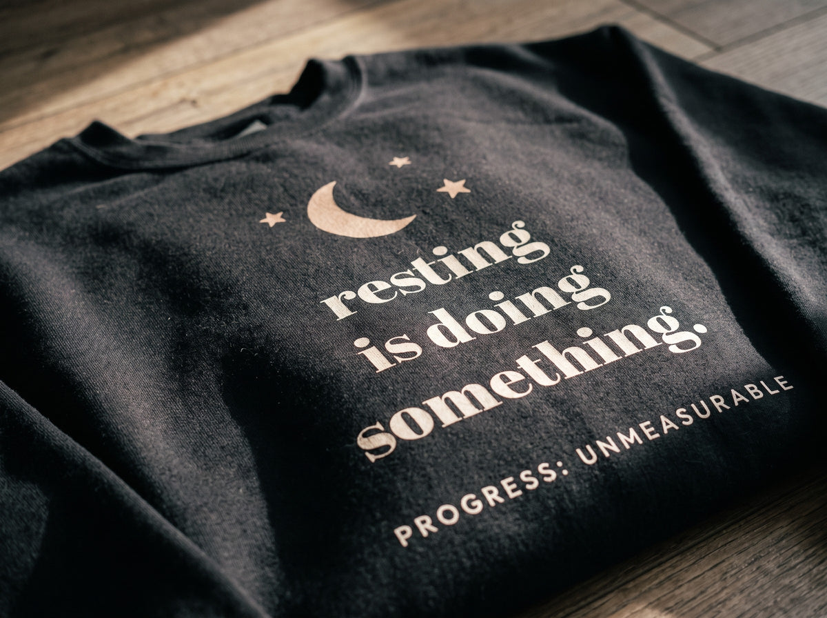 Resting Is Doing Something Crewneck, the print up close on black fleece: a moon and three stars, resting is doing something. and PROGRESS: UNMEASURABLE.