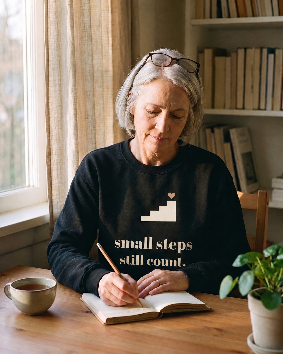 Small Steps Still Count crewneck in Black, worn by a woman writing in a notebook at a desk by a window. Printed on the chest: a four-step staircase with a small heart on top and the words small steps still count.
