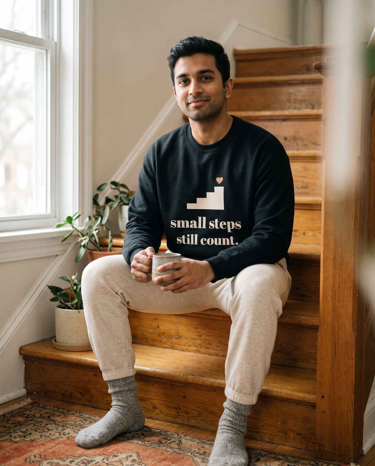Small Steps Still Count crewneck in Black, worn by a model sitting on a wooden staircase with a mug. Printed on the chest: small steps still count, with a staircase and a small heart.