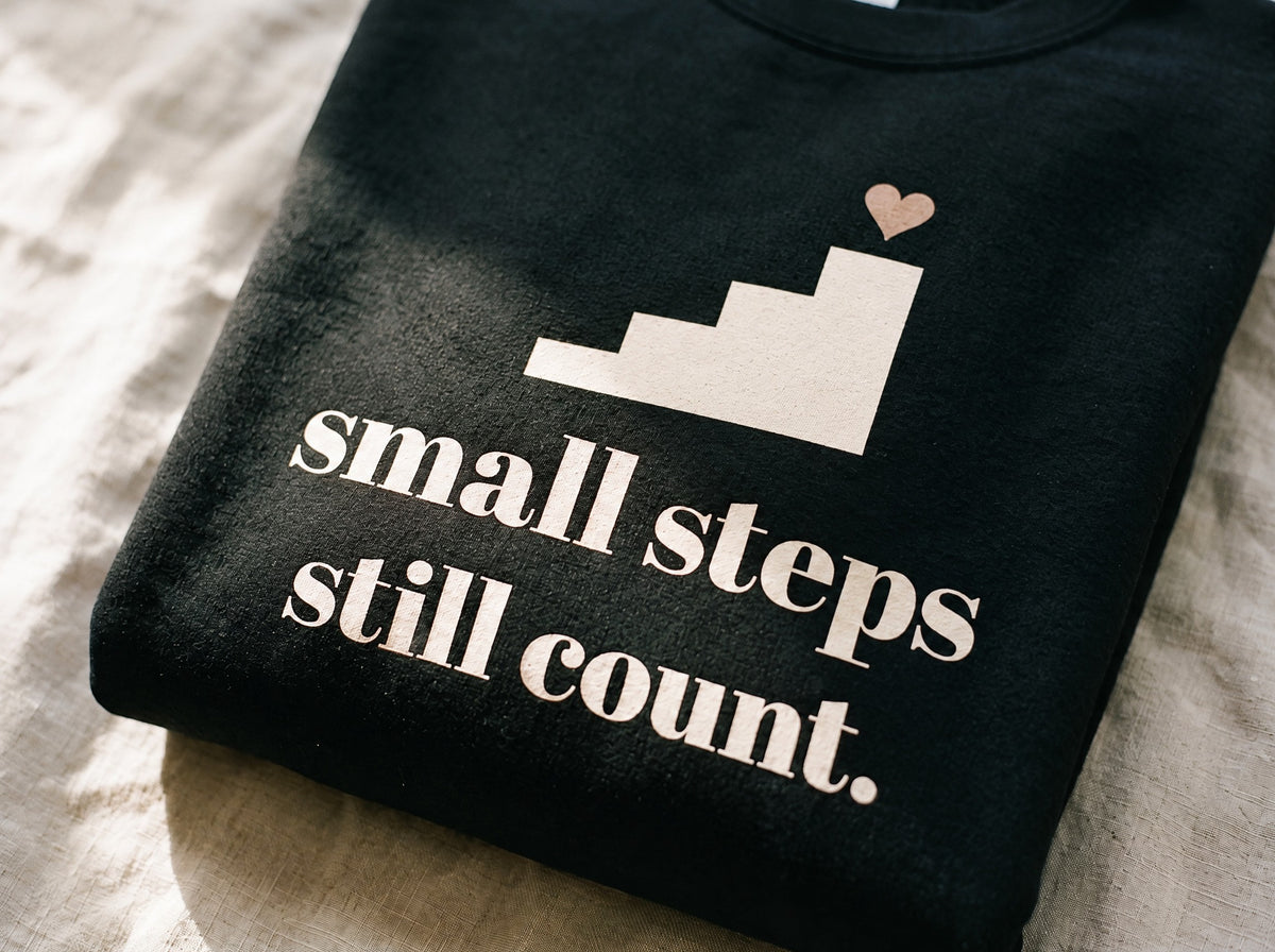 Small Steps Still Count Crewneck, the print up close on black fleece: a four-step staircase with a small heart on top and small steps still count.