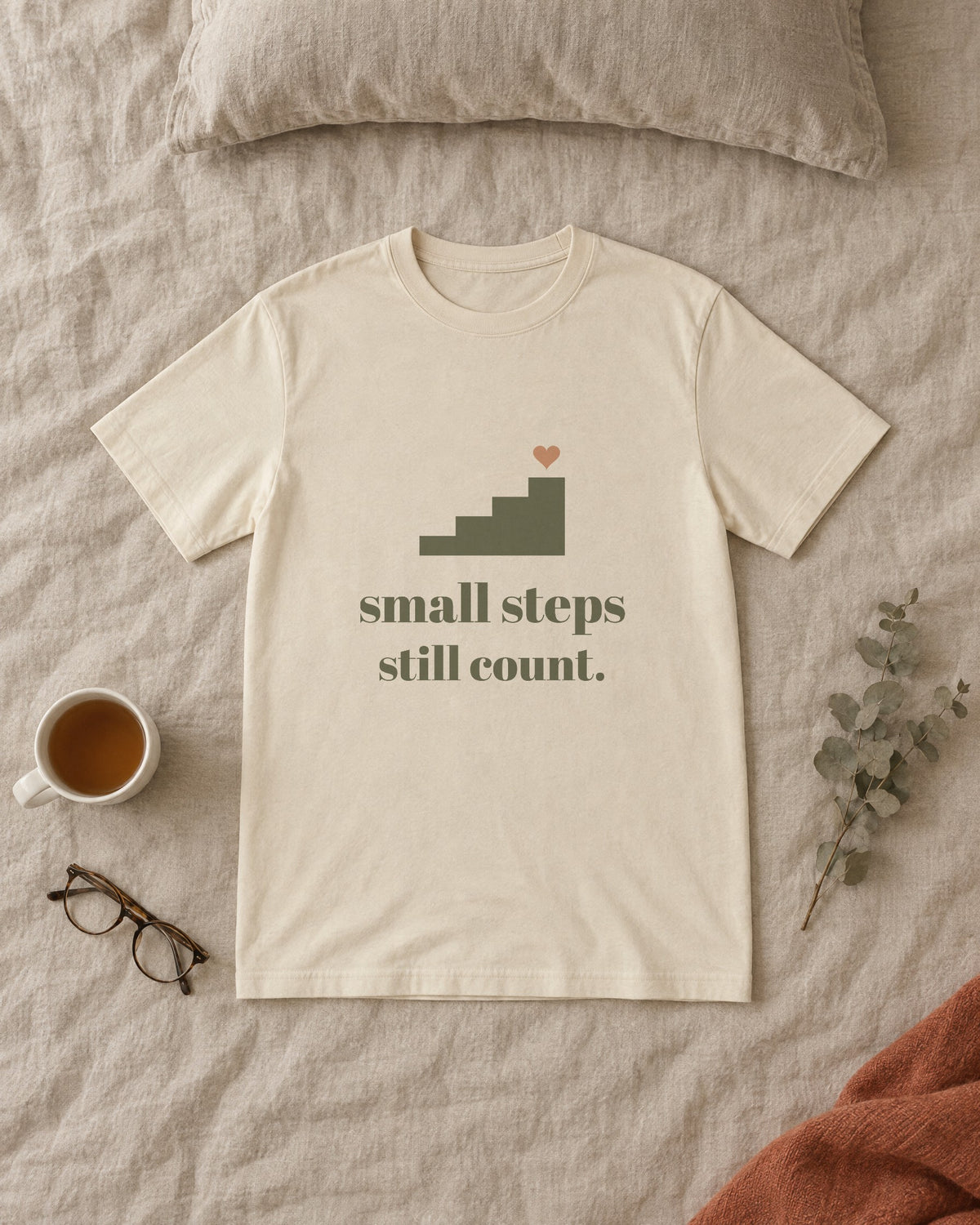 Small Steps Still Count Tee, in soft cream, laid flat on a made bed