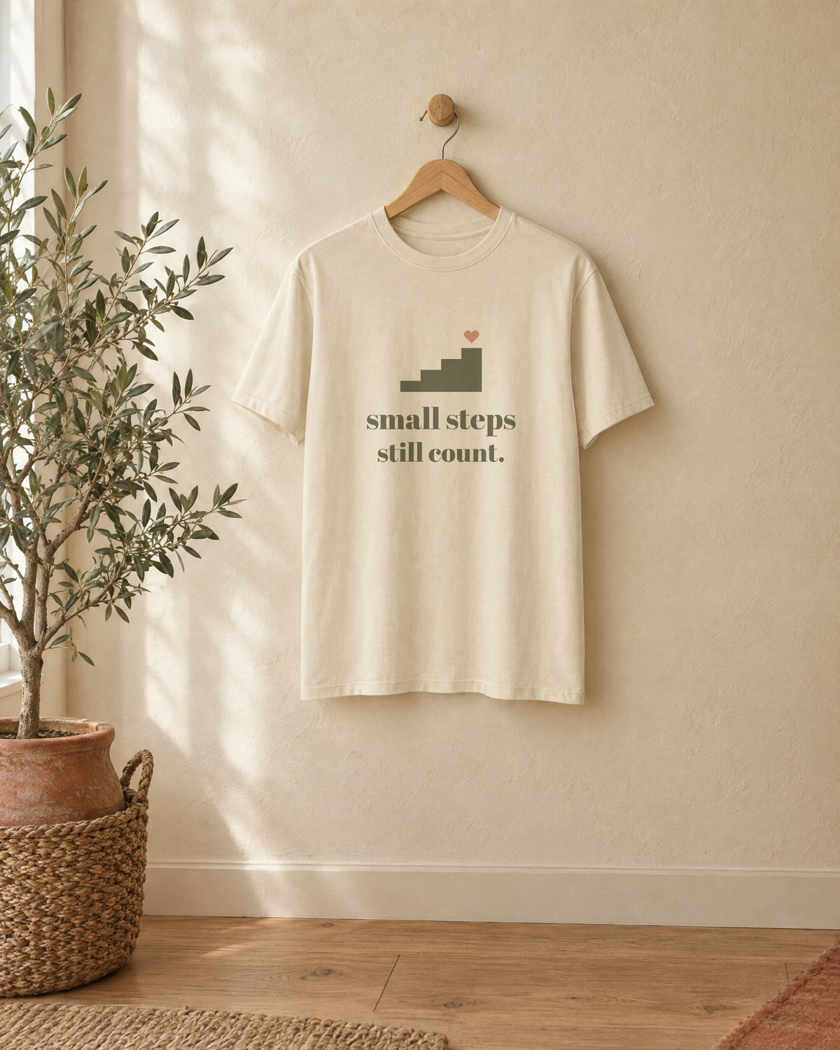 Small Steps Still Count Tee, in soft cream, on a wooden hanger against a cream wall