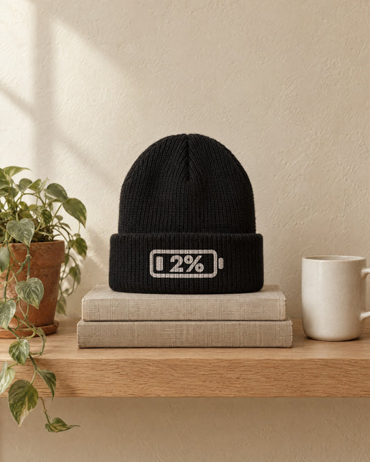 Social Battery Beanie in Black, sitting on a stack of books on an oak shelf. Embroidered on the cuff: a battery at 2%.