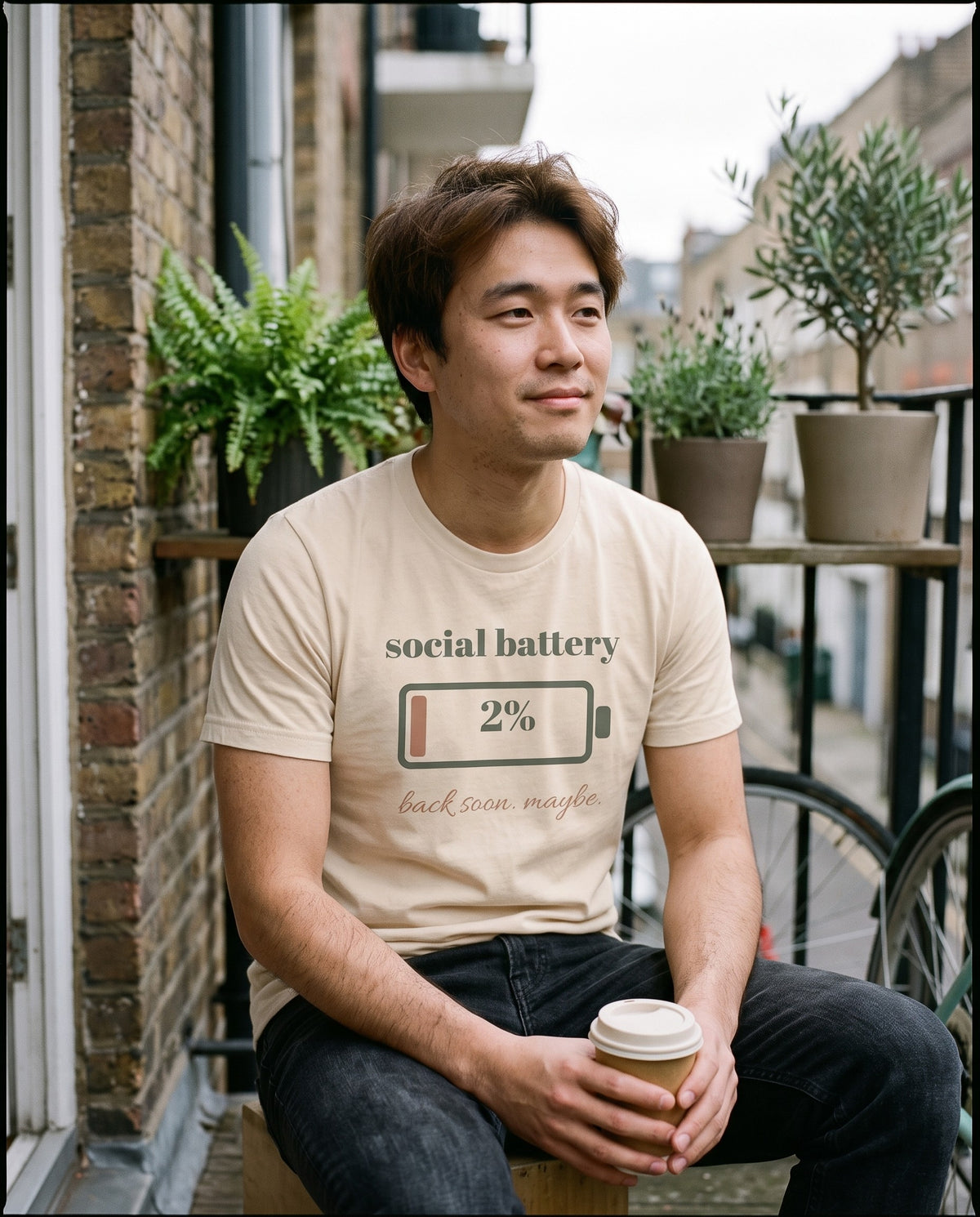 Social Battery Tee, worn with a takeaway coffee on a city balcony