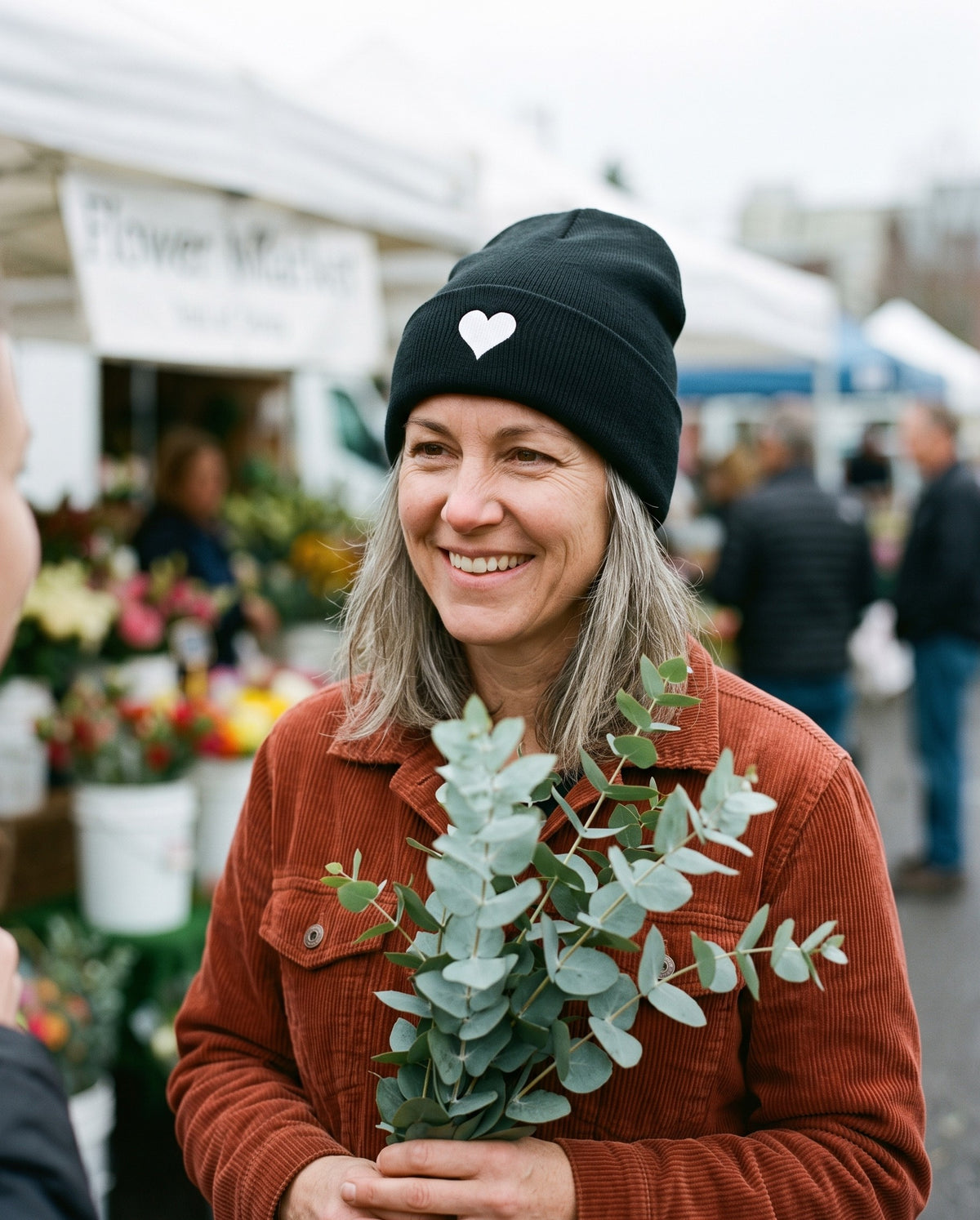 Soft Heart Beanie in Black, worn by a woman holding eucalyptus at a flower market. Embroidered on the cuff: a small white heart.