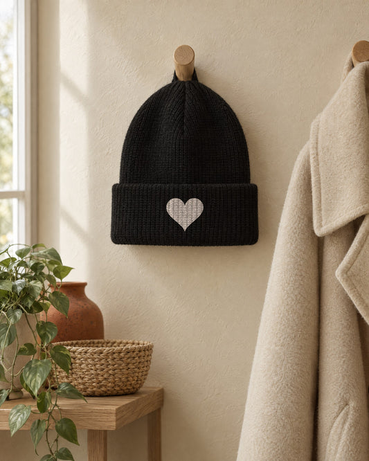 Soft Heart Beanie in Black, hanging on a wooden wall peg by a window. Embroidered on the cuff: a small heart.