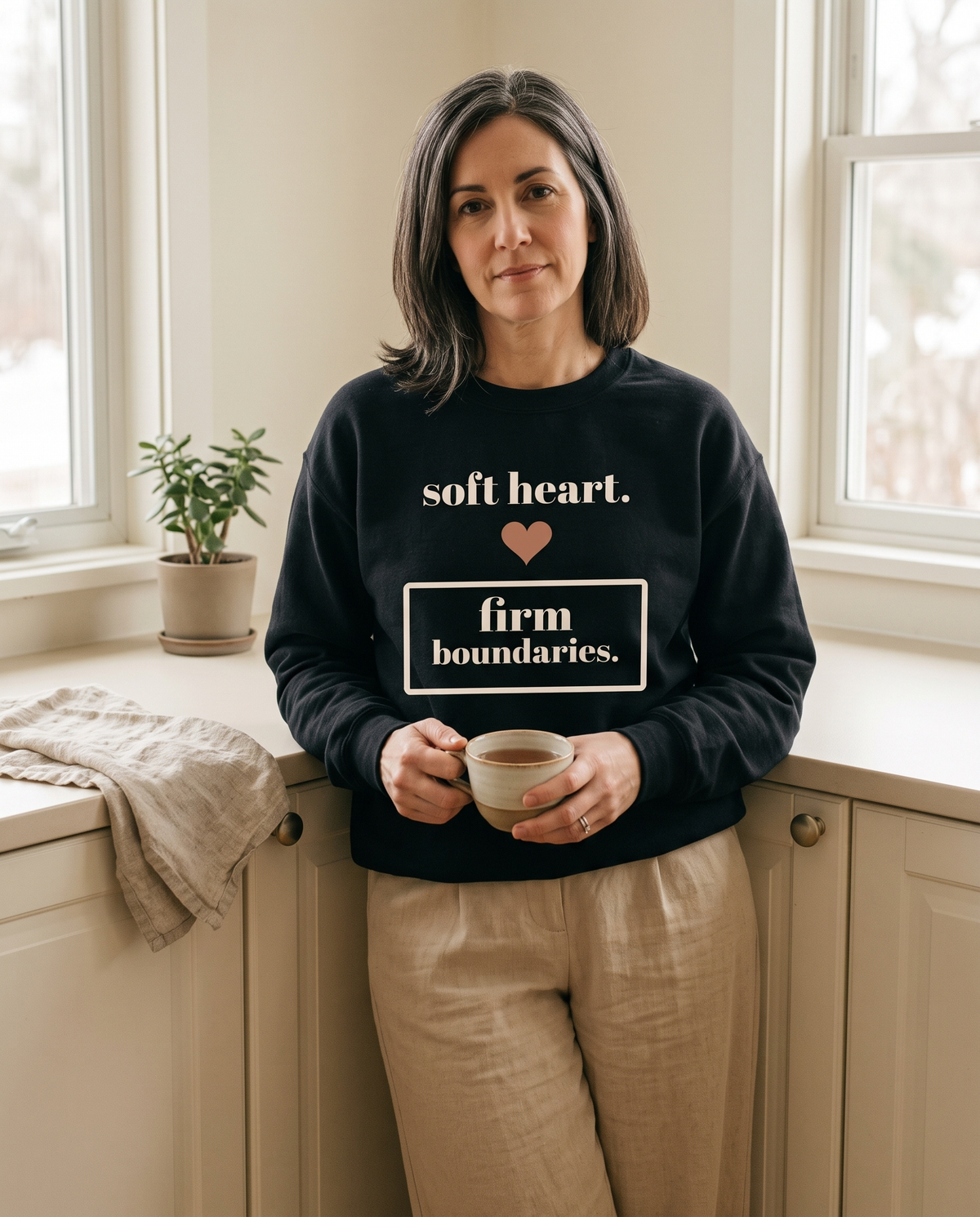 Soft Heart, Firm Boundaries crewneck in Black, worn by a model leaning on a kitchen counter with a cup of tea. Printed on the chest: soft heart, firm boundaries, with a small heart.