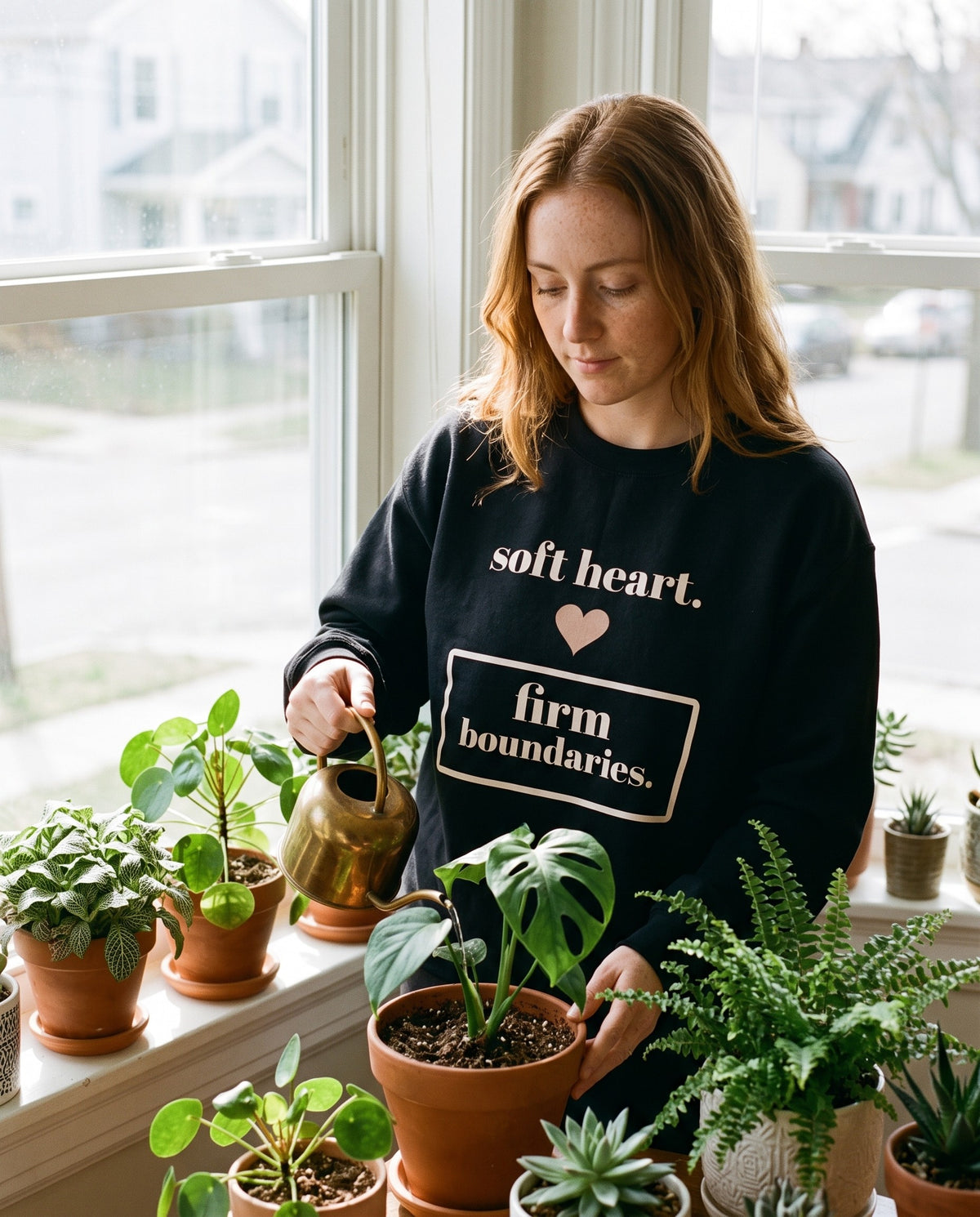 Soft Heart, Firm Boundaries crewneck in Black, worn by a woman watering plants on a sunny windowsill. Printed on the chest: soft heart., a small heart and firm boundaries. inside a box.